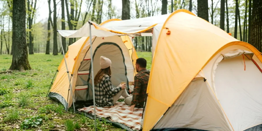 Two people sitting at the entrance of a yellow and white tent in a woodland clearing